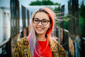 A young woman with multi-colored hair and glasses smiles at the camera.
